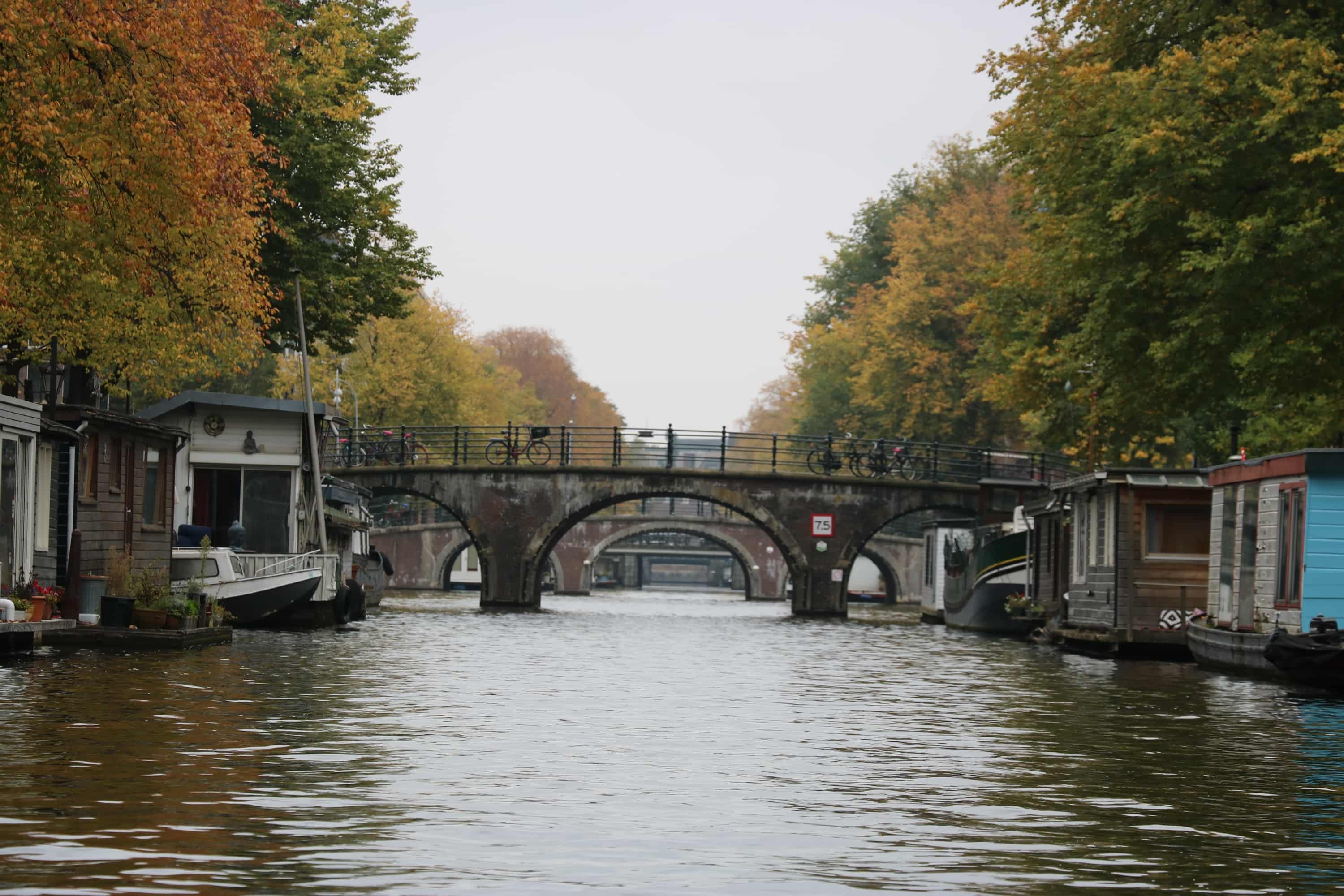 A grey sky over the murky waters of one of Amsterdam's canals. An arched bridge runs across the canal in the background.