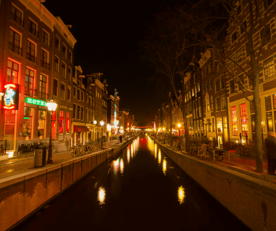 On of Amsterdam's famous canals runs through the middle of the image. On either side are traditional Dutch buildings lit up with red lights to symbolise the Red Light district.