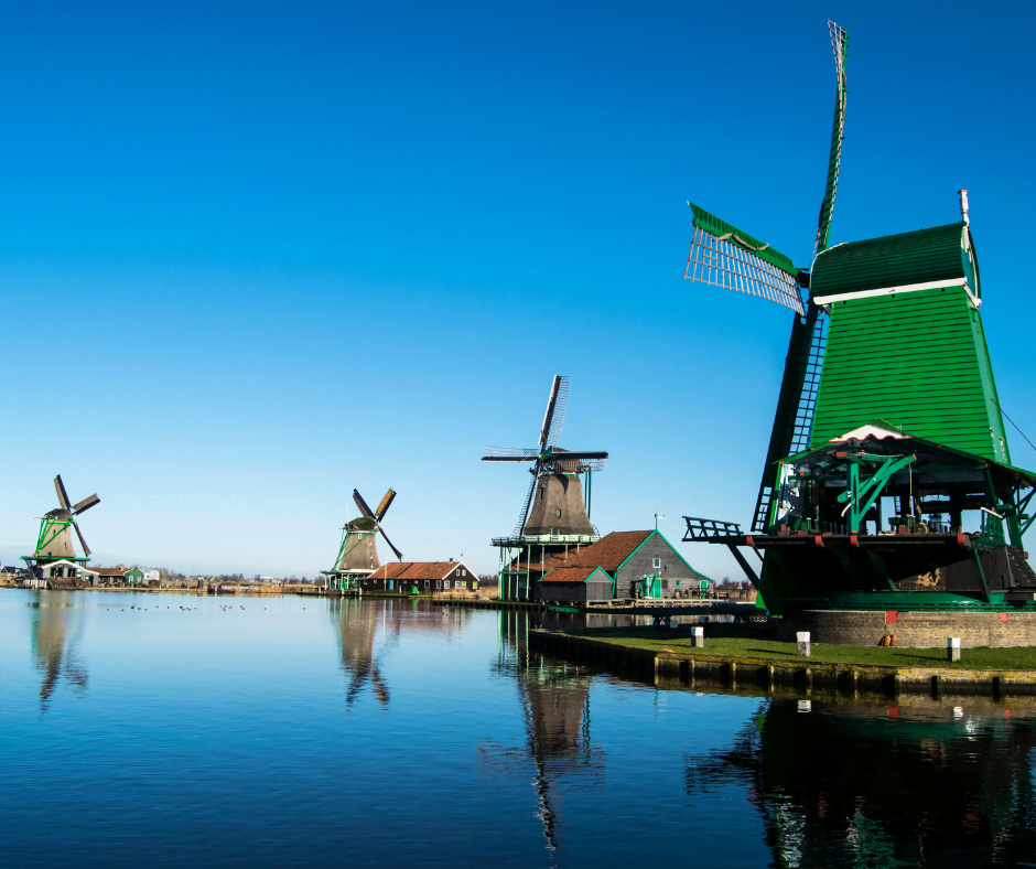 A mirror-still river with green windmills lining the banks. The sky is blue and reflected in the water. 