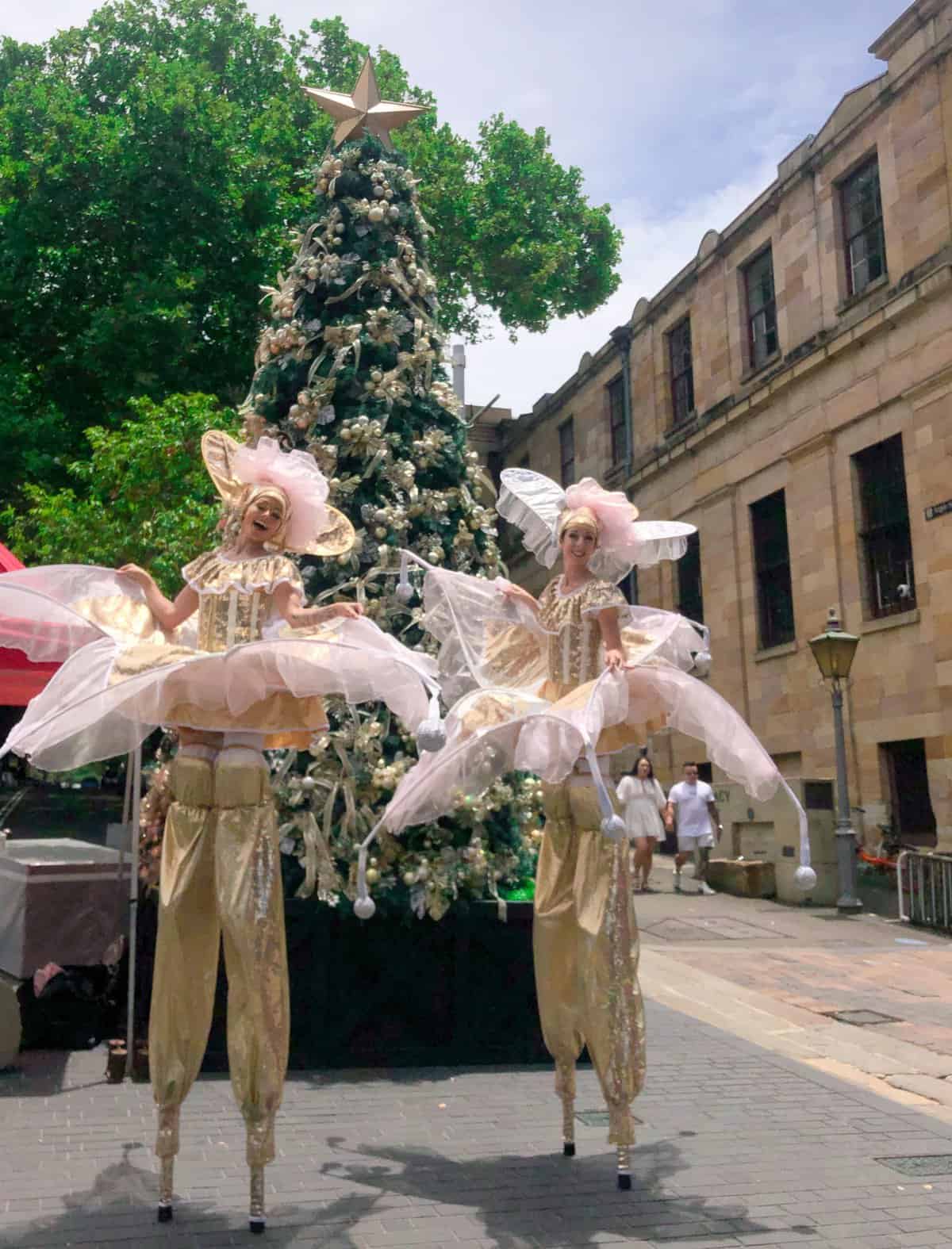 Two performers on stilts posing in front of a Christmas Tree outdoors at The Rocks Christmas Market