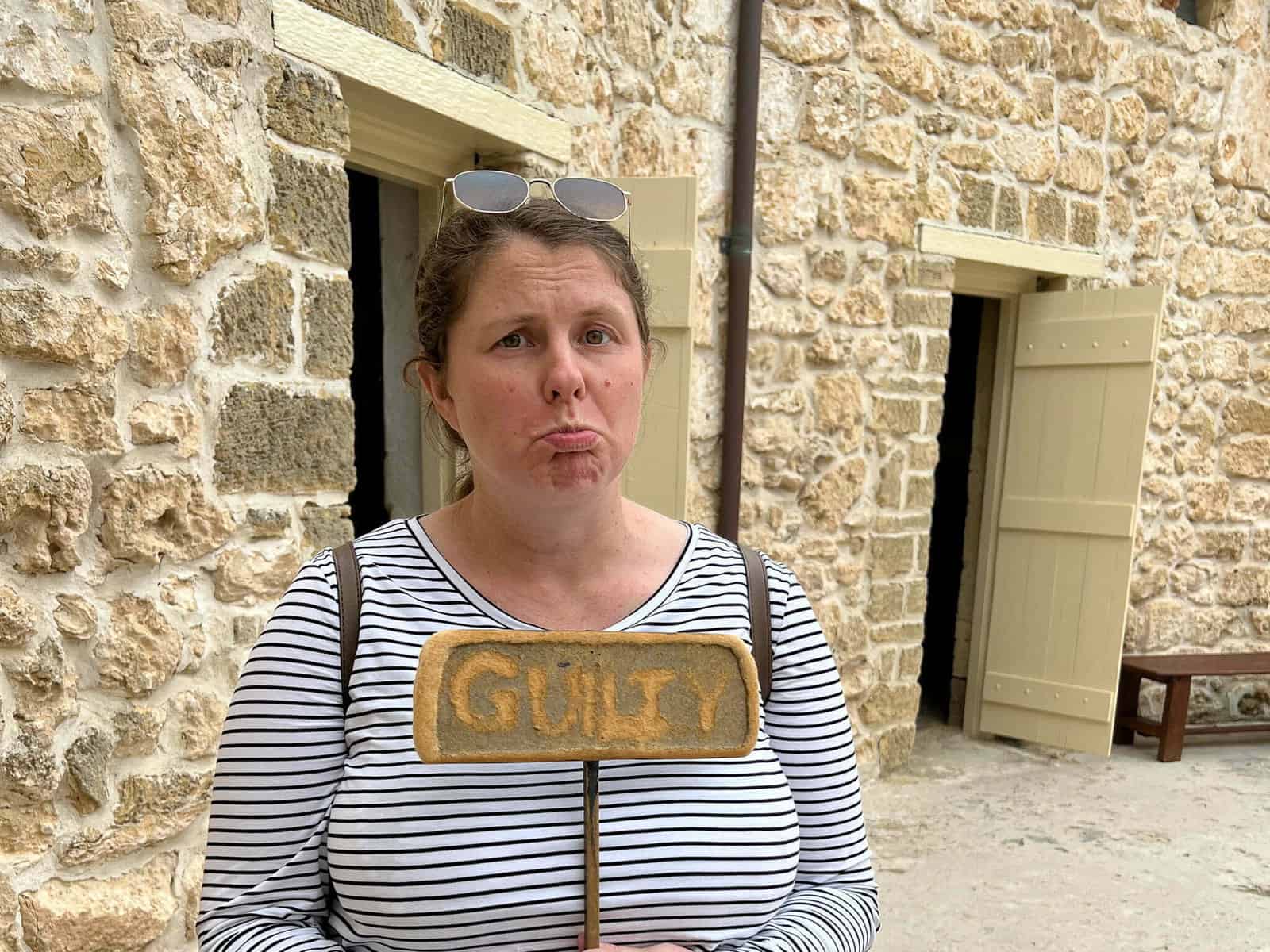 Emma stands in the centre of the Fremantle Round House courtyard holding a sign saying "Guilty". She is making a sad face in jest.