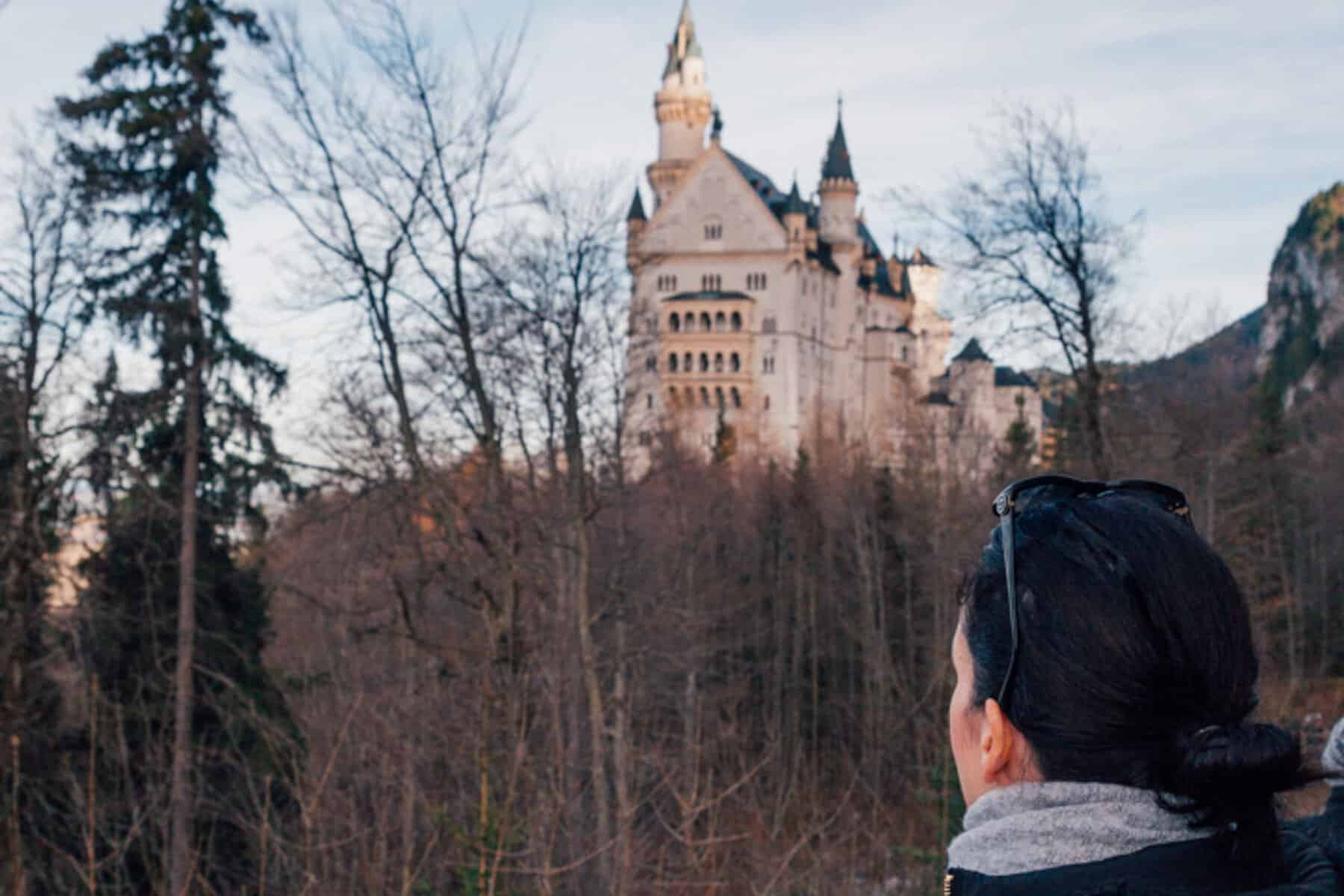 Lucia is facing the castle looking up through the forest to the castle on the hill. She has dark hair tied in a bun and is wearing sunglasses on her head.