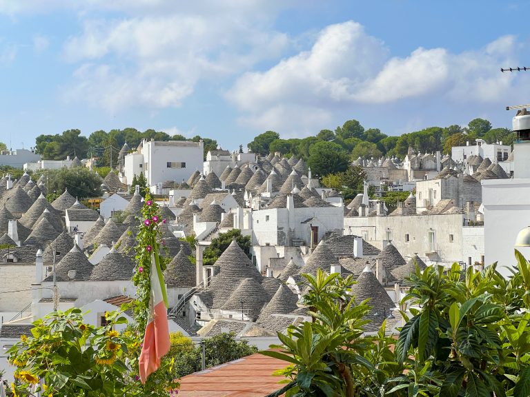 Colourful hillside view of traditional Italian trulli houses with conical roofs in Alberobello, Puglia, Italy, under a bright blue sky with puffy clouds.
